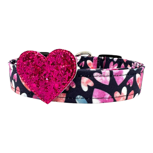 Valentine’s Day dog collar with pink and blush watercolor heart pattern on a dark background with removable glitter heart embellishment, shown flat with buckle and hardware visible.