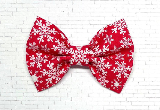 Red dog bow tie with a white snowflake pattern, displayed on a white background. The festive bow tie features elastic loops on the back for easy attachment to a dog collar.