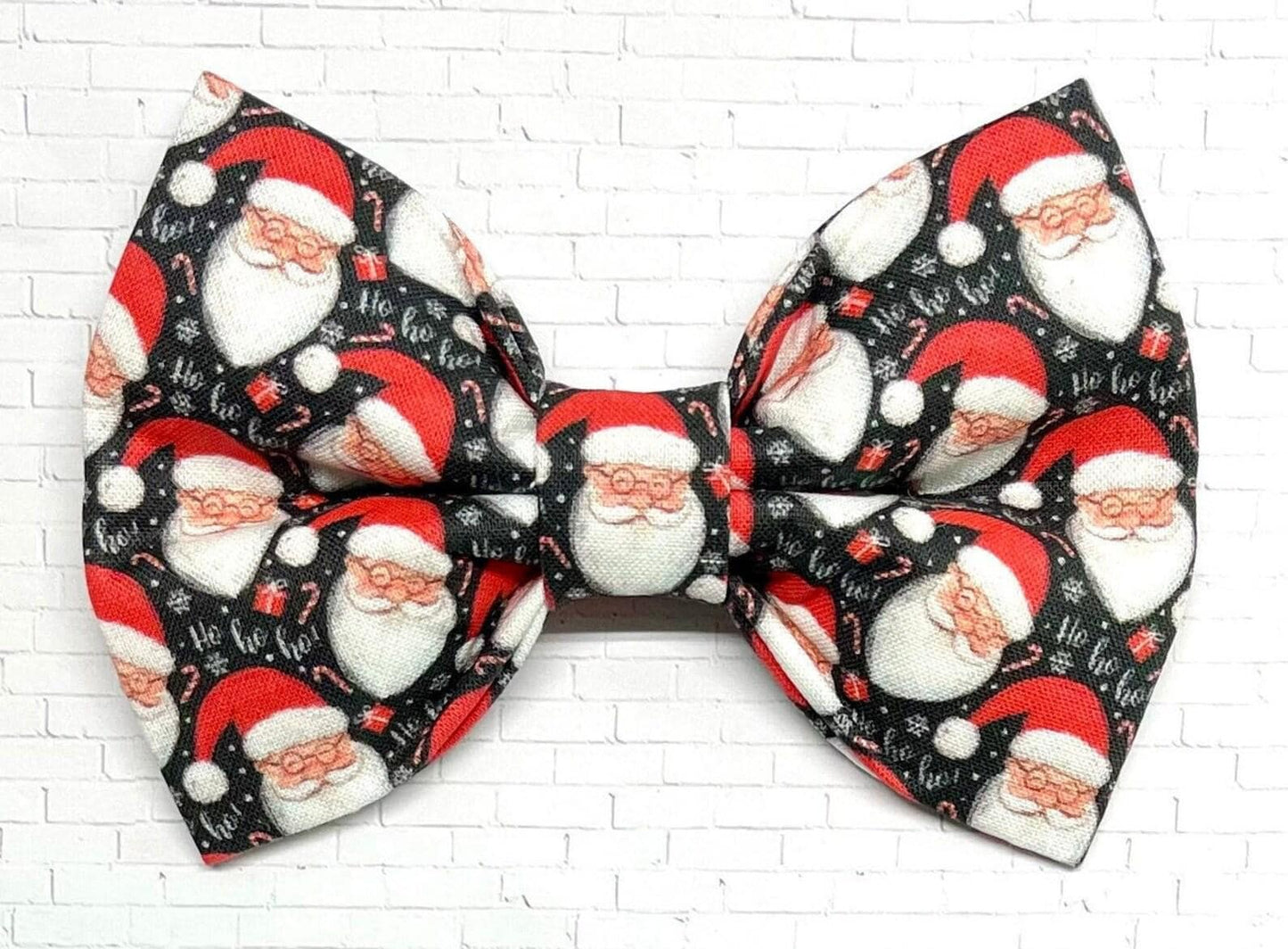 Black dog bow tie with a festive pattern of cheerful Santa faces in red and white, shown on a white background. The handmade bow tie includes a loop on the back for easy attachment to a collar.