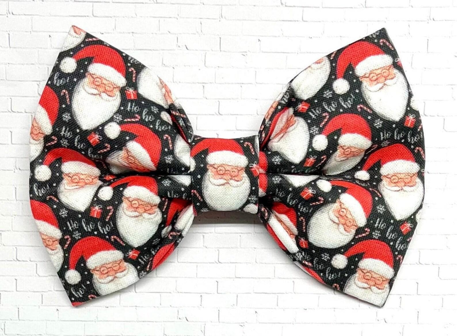 Black dog bow tie with a festive pattern of cheerful Santa faces in red and white, shown on a white background. The handmade bow tie includes a loop on the back for easy attachment to a collar.