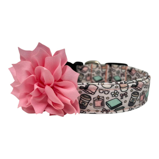 Pale pink dog collar with whimsical illustrations of books, coffee cups, candles, daisies, bows, and glasses, paired with a removable flower attached by an elastic loop.