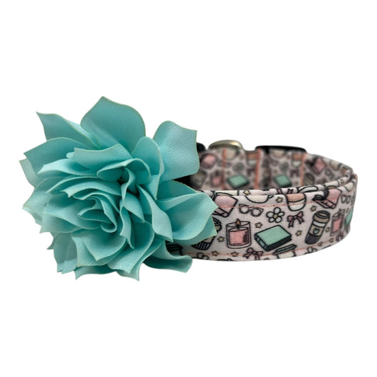 Pale pink dog collar with whimsical illustrations of books, coffee cups, candles, daisies, bows, and glasses, paired with a removable flower attached by an elastic loop.