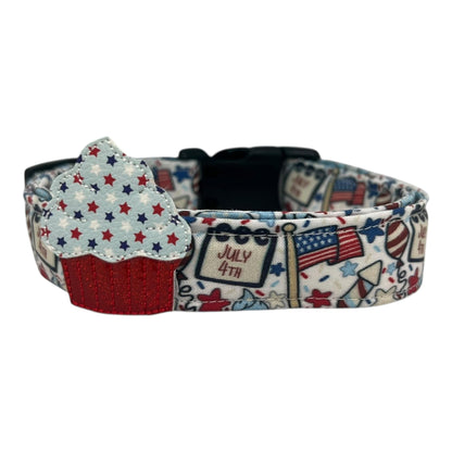 Light-colored 4th of July dog collar featuring festive red, white, and blue designs like American flags, balloons, stars, and rockets, with a removable patriotic embellishment attached by an elastic loop.