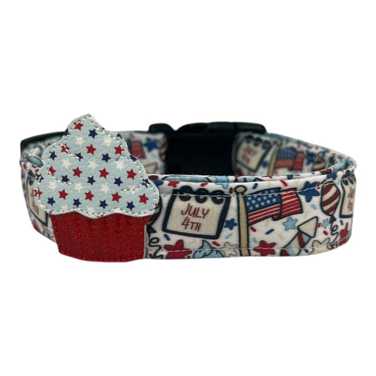 Light-colored 4th of July dog collar featuring festive red, white, and blue designs like American flags, balloons, stars, and rockets, with a removable patriotic embellishment attached by an elastic loop.
