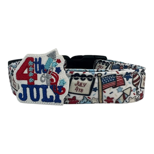 Light-colored 4th of July dog collar featuring festive red, white, and blue designs like American flags, balloons, stars, and rockets, with a removable patriotic embellishment attached by an elastic loop.