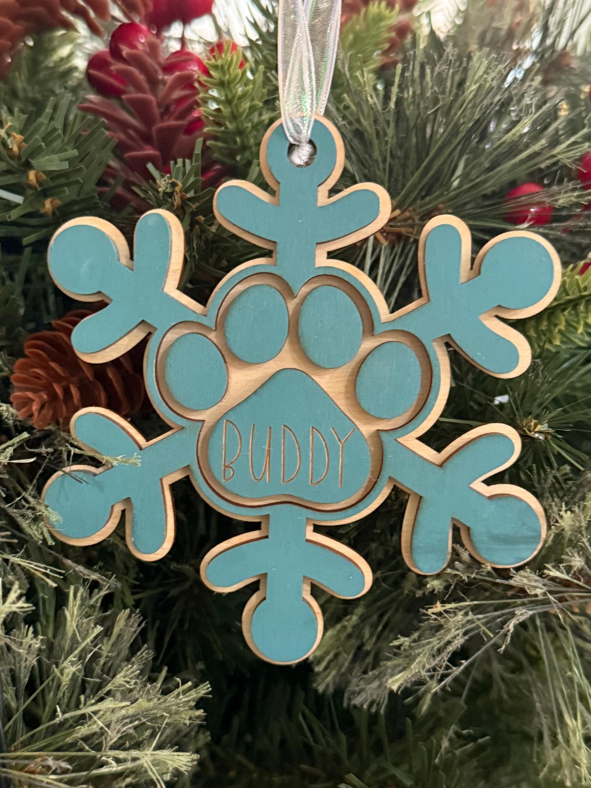 Personalized wooden dog paw snowflake ornament in blue with custom engraved pet name and white hanging ribbon, displayed as holiday décor