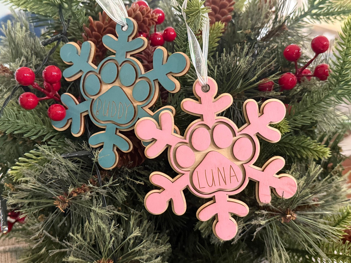 Personalized wooden dog paw snowflake ornament in pink or blue with custom engraved pet name and white hanging ribbon, displayed as holiday décor