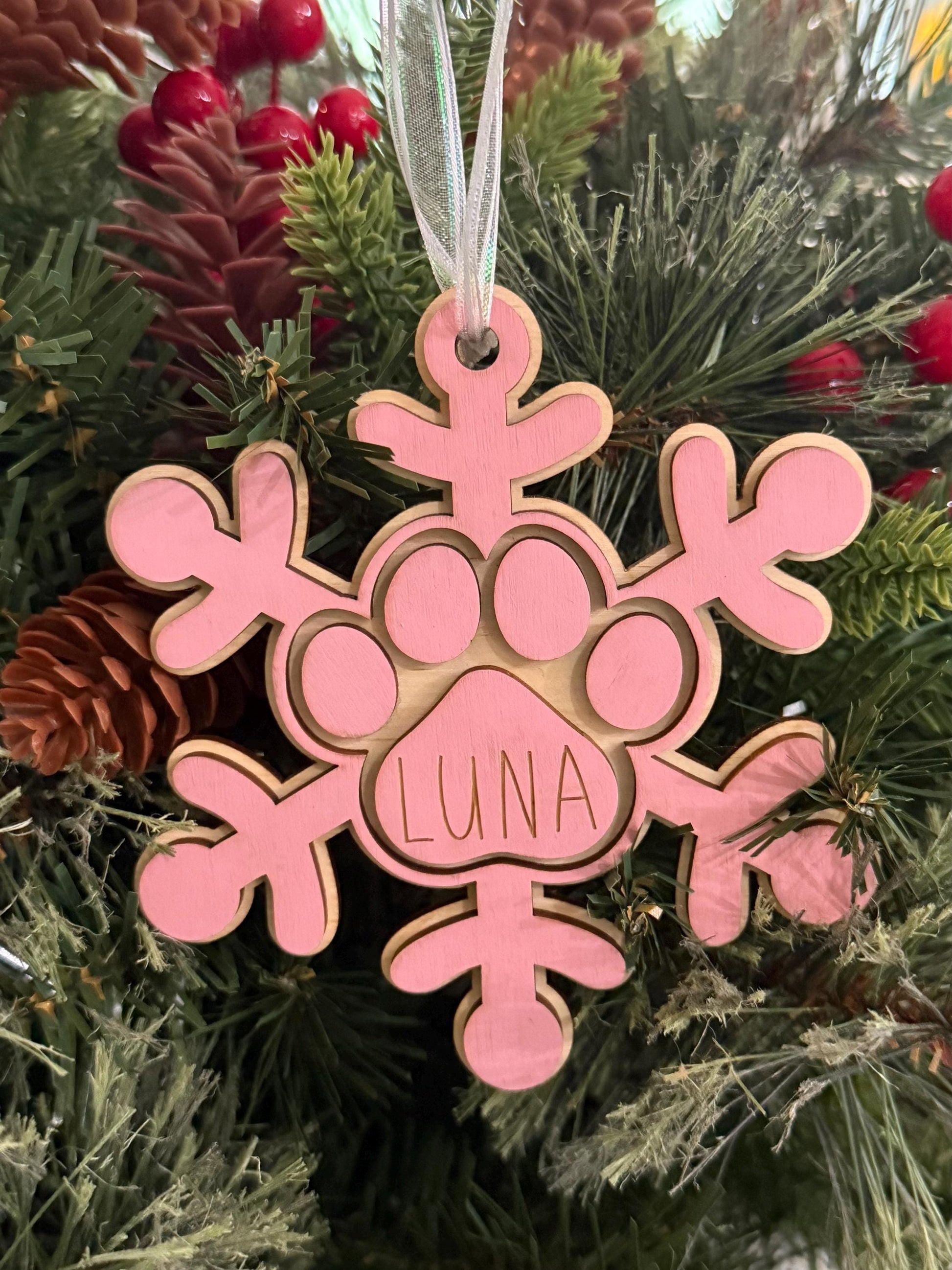 Personalized wooden dog paw snowflake ornament in pink with custom engraved pet name and white hanging ribbon, displayed as holiday décor