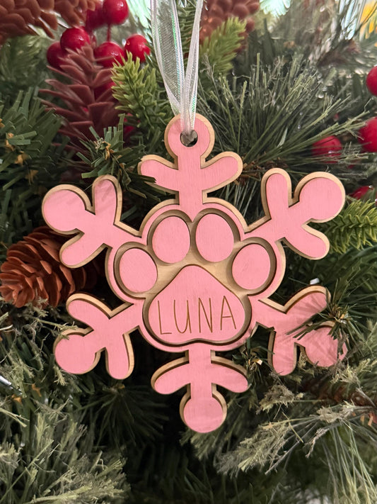 Personalized wooden dog paw snowflake ornament in pink with custom engraved pet name and white hanging ribbon, displayed as holiday décor