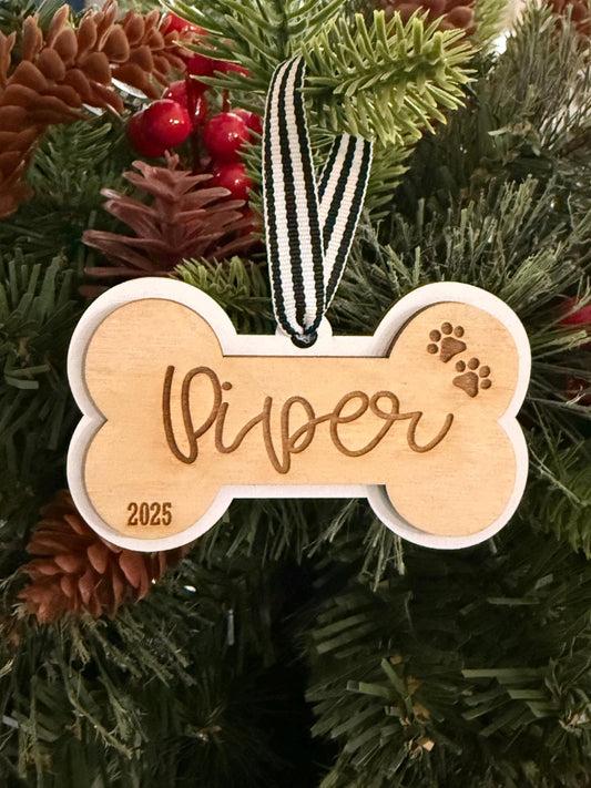 Personalized wooden dog bone Christmas ornament engraved with dog’s name, year, and paw prints, finished with a white border and striped hanging ribbon